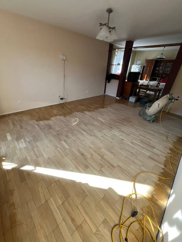 Cyklinowanie parkietu w trakcie realizacji. Część podłogi już wycyklinowana, widoczna maszyna do cyklinowania i kabel zasilający. Jasne drewno, naturalne światło.