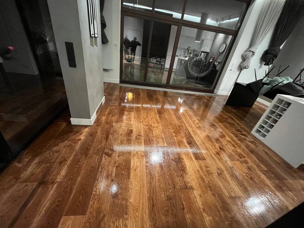 Błyszcząca, świeżo polakierowana podłoga z desek w odcieniach brązu, odbijająca światło. Widoczne duże okno przesuwne i fragmenty wyposażenia wnętrza.