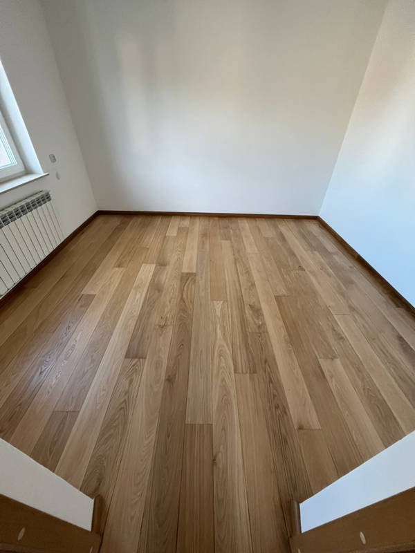 Jasna, drewniana podłoga w pokoju na poddaszu, widok z dołu schodów. Naturalne oświetlenie podkreśla strukturę drewna i białe ściany.