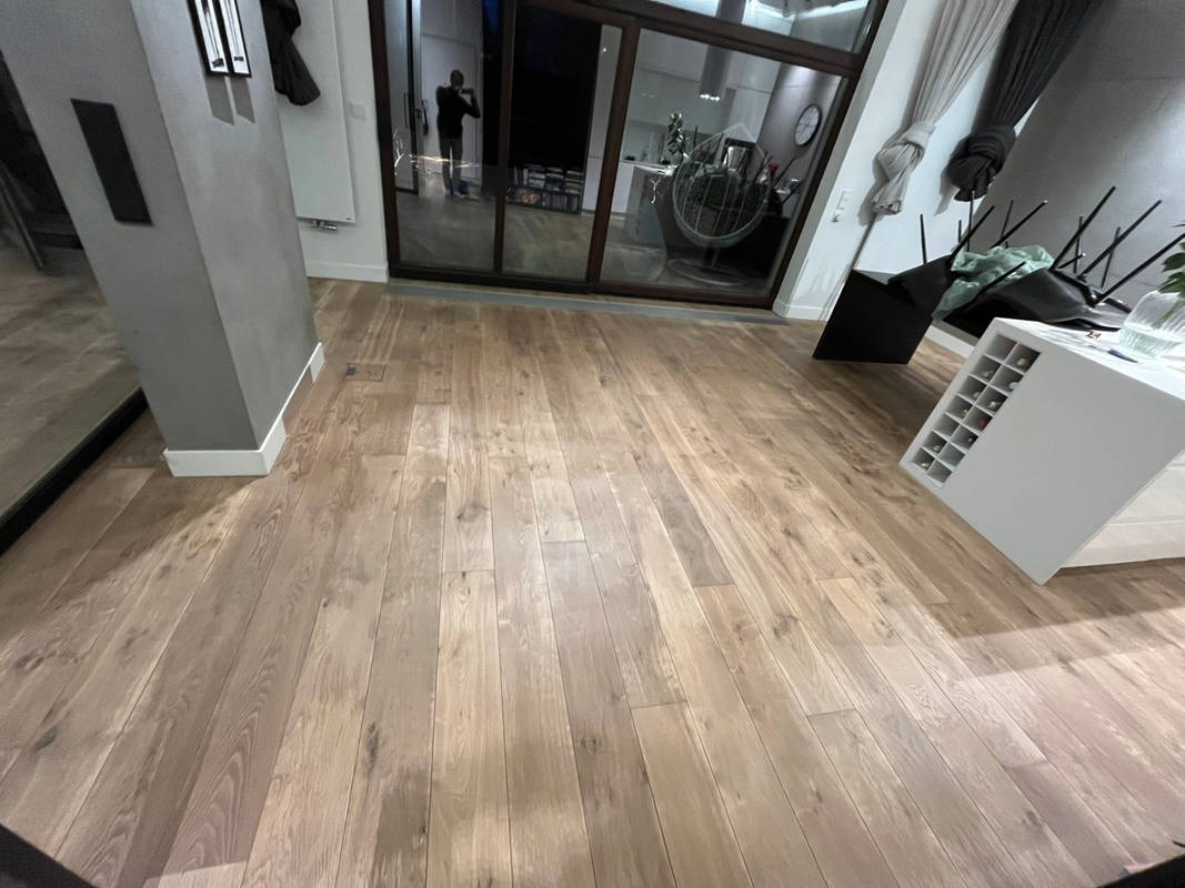 Jasna, drewniana podłoga ułożona w salonie z dużym oknem przesuwnym. Widoczny fragment kolumny i mebli. Naturalne oświetlenie podkreśla strukturę drewna.