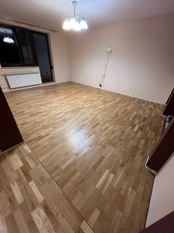 Jasny parkiet ułożony w jodełkę w pustym pokoju z grzejnikiem i oknem balkonowym. Widoczne ślady po listwach przypodłogowych i gniazdko elektryczne na ścianie.