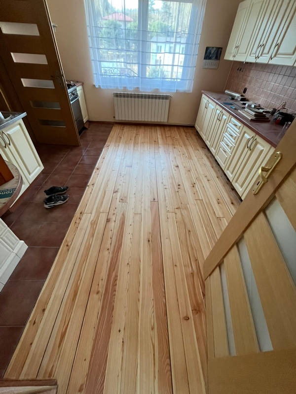 Jasna, drewniana podłoga z widocznymi słojami w kuchni, świeżo po ułożeniu. W tle okno z firanką i grzejnik. Częściowo widoczne meble kuchenne.