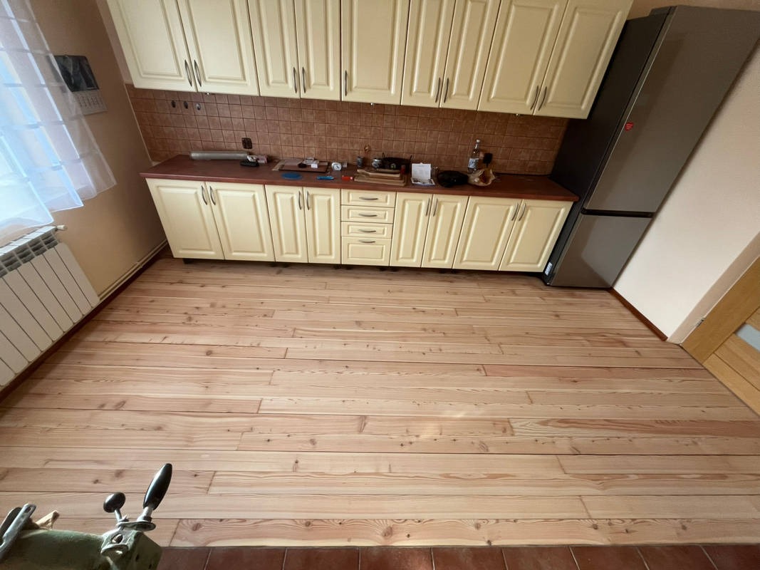 Jasna, drewniana podłoga w kuchni z kremowymi szafkami. Widoczny fragment maszyny do szycia w lewym dolnym rogu kadru. Ukończona realizacja.