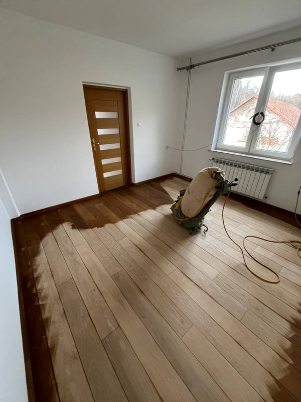 Cyklinowanie parkietu w Otwocku w trakcie realizacji. Widoczna maszyna do cyklinowania i fragment podłogi po obróbce, ukazujący różnicę w kolorze drewna.