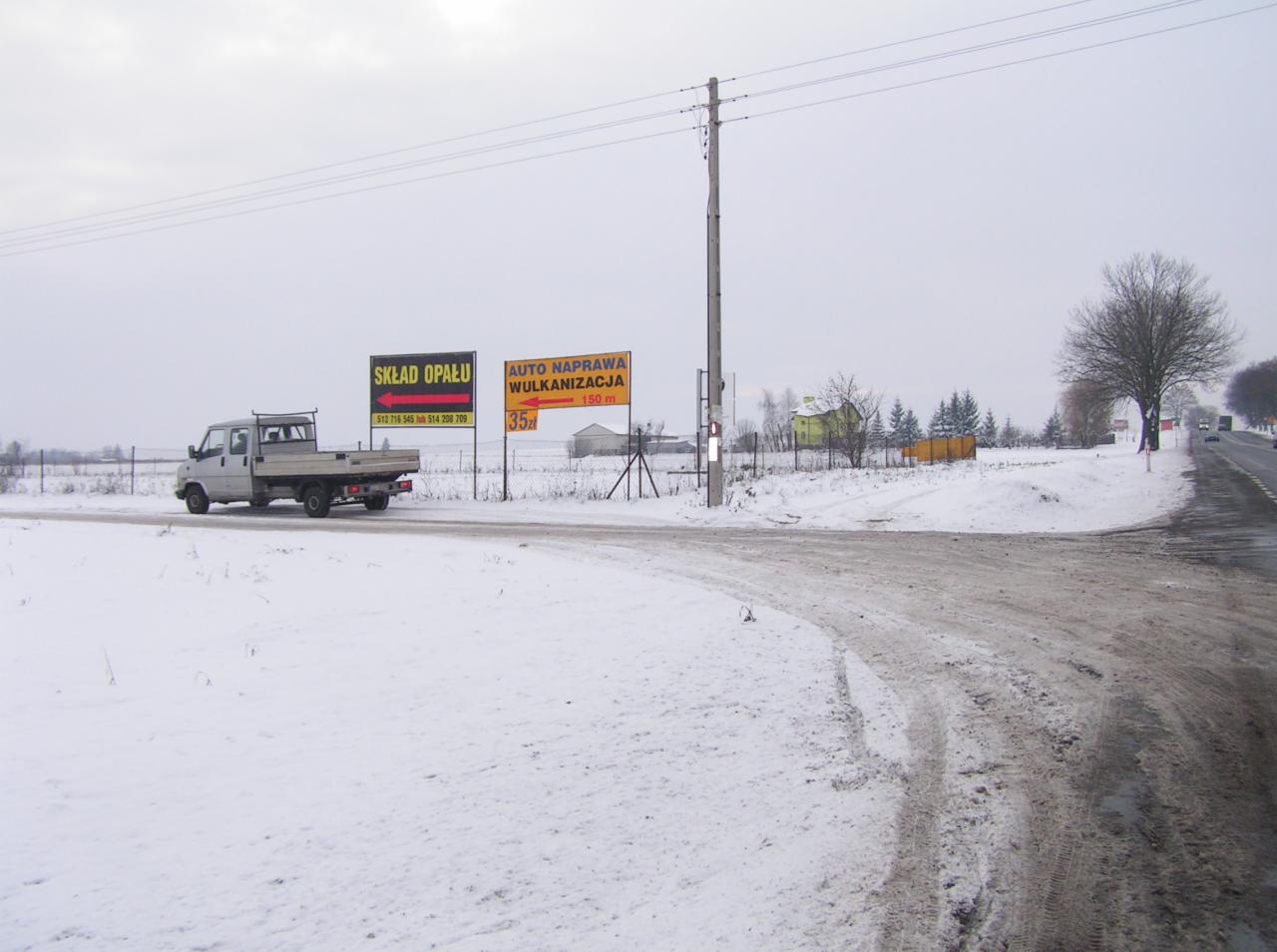 Zimowy krajobraz z samochodem dostawczym na tle tablic reklamowych składu opału i auto naprawy.