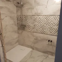 Montaż kabiny prysznicowej z białego marmuru z deszczownicą i wzorzystą mozaiką w strefie mokrej, widoczne nieukończone wykończenia ściany.