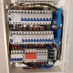 Rozdzielnica elektryczna z bezpiecznikami Schrack i Schelinger, widoczne przewody w kolorach niebieskim, czarnym, brązowym, żółto-zielonym i czerwonym, zamontowana w białej obudowie.