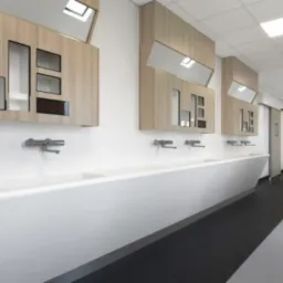 Długi, publiczny korytarz z umywalkami, drewnianymi szafkami z otwartymi przestrzeniami i czarno-białą podłogą, oświetlony przez kwadratowe panele sufitowe.