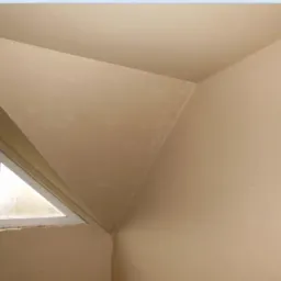 Gładko otynkowane poddasze z trójkątnym oknem. Ściany w jasnym, neutralnym odcieniu, świeżo po tynkowaniu. Widoczne przejścia między ścianami i sufitem.
