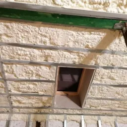 Ocieplanie poddasza pianką PUR między metalowymi profilami, widoczny fragment okna dachowego i drewniana podpora konstrukcyjna.