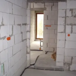 Wnętrze w trakcie instalacji elektrycznej: widoczne puszki instalacyjne w kolorze pomarańczowym, przewody elektryczne prowadzone po ścianach z bloczków betonowych, czarne rury instalacyjne przy...