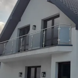 Balkon z nowoczesną szklaną balustradą na tle białej elewacji domu z ciemną dachówką i oknami.