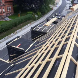 Konstrukcja drewniana dachu z membraną Ecotec i logotypem Monier, widok z góry na przygotowany do wymiany dach, w tle zabudowa miejska i zieleń.