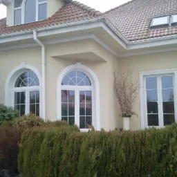 Jasnożółty dom jednorodzinny z dwoma oknami łukowymi i jednym prostokątnym, białe ramy okienne, czerwona dachówka, zadbany żywopłot przed budynkiem i dekoracja z gałęzi w wazonie.