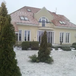 Jasny, dwukondygnacyjny dom jednorodzinny z balkonem, czerwonym dachem pokrytym śniegiem i dużymi oknami, otoczony zimowym ogrodem z iglakami.