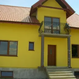 Żółty dom jednorodzinny z balkonem i brązowymi oknami, widoczne schody wejściowe i fragment fundamentu.