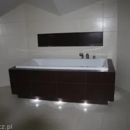 Wykończona łazienka na poddaszu: zabudowana wanna z brązowymi frontami podświetlona od dołu, jasne płytki na ścianach i podłodze, ciemny pas dekoracyjny na ścianie.