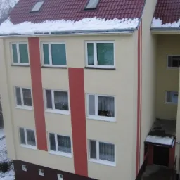 Wielopiętrowy budynek po renowacji z beżową elewacją, czerwonymi pasami pionowymi i dachem z czerwonej dachówki, częściowo pokrytym śniegiem. Widoczne okna dachowe i śnieg na parapetach. Ujęcie...