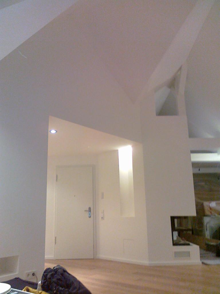 Wnętrze po remoncie: białe ściany z zabudową z płyt gipsowo-kartonowych, widoczne wykończenia gładzią i malowaniem, drzwi wejściowe po lewej stronie, fragment podłogi i niedokończony kominek po...