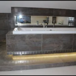 Wykończona łazienka z wanną obłożoną brązowymi płytkami imitującymi kamień, podświetlaną listwą LED u podstawy i lustrami wnękowymi nad wanną.