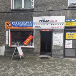 Fasada budynku z kilkoma szyldami firm: salon fryzjerski dla zwierząt Psi Zakątek, szkoła językowa Pegaz Direct, biuro rachunkowe Koan oraz reklama kredytów z napisem 'Kredyty' na czerwonym tle...