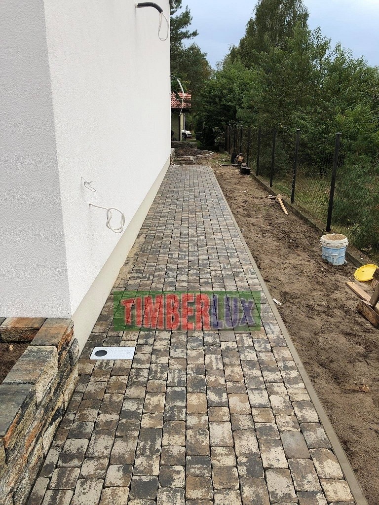 Ułożona kostka brukowa w odcieniach szarości i beżu, przylegająca do białej ściany budynku z widocznymi elementami instalacji zewnętrznej, z odpływem liniowym i fragmentem murku z kamienia...