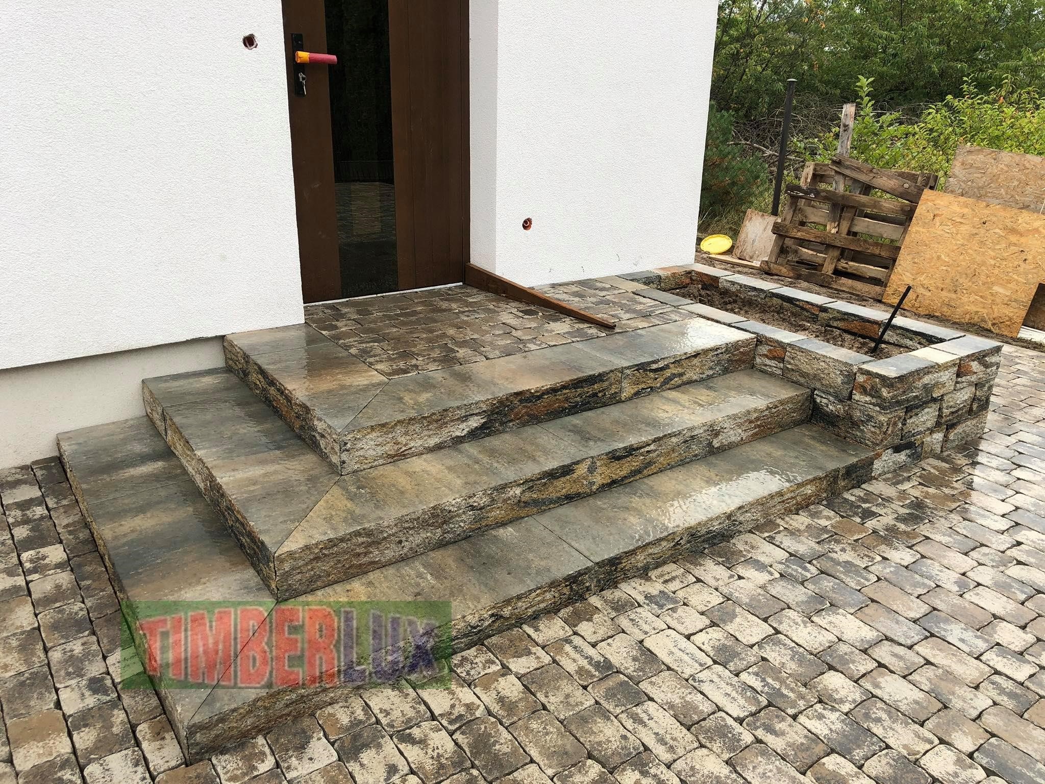 Trzy stopnie wejściowe wykonane z szarej kostki o nieregularnej fakturze, prowadzące do drzwi budynku; obok widoczny fragment murku z tej samej kostki i nawierzchnia z małych, prostokątnych kostek...