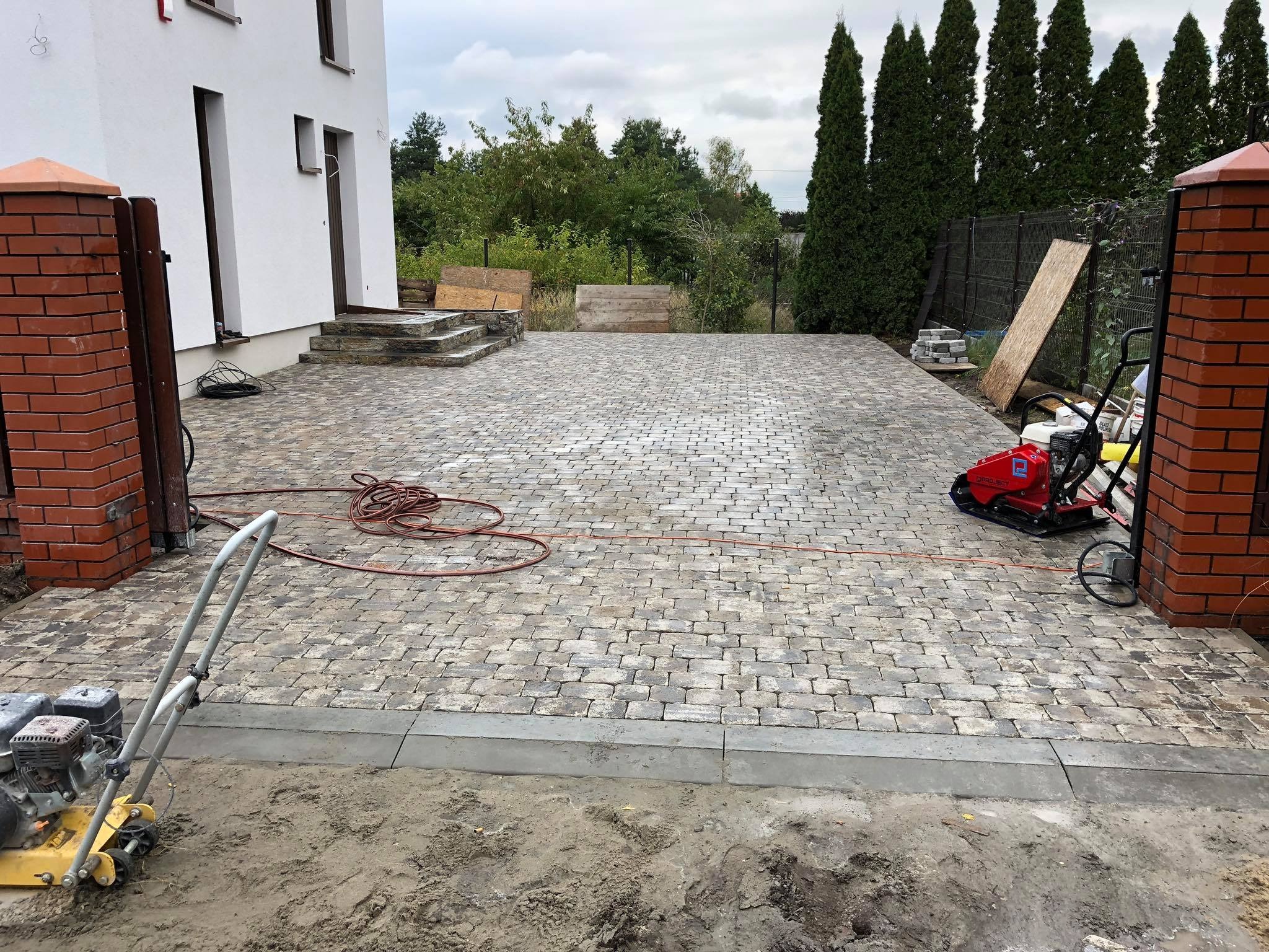 Ułożona kostka brukowa na podjeździe z czerwonymi słupkami bramy, widoczne zagęszczarki w tle i na pierwszym planie, oraz obrzeża betonowe oddzielające nawierzchnię od ziemi.