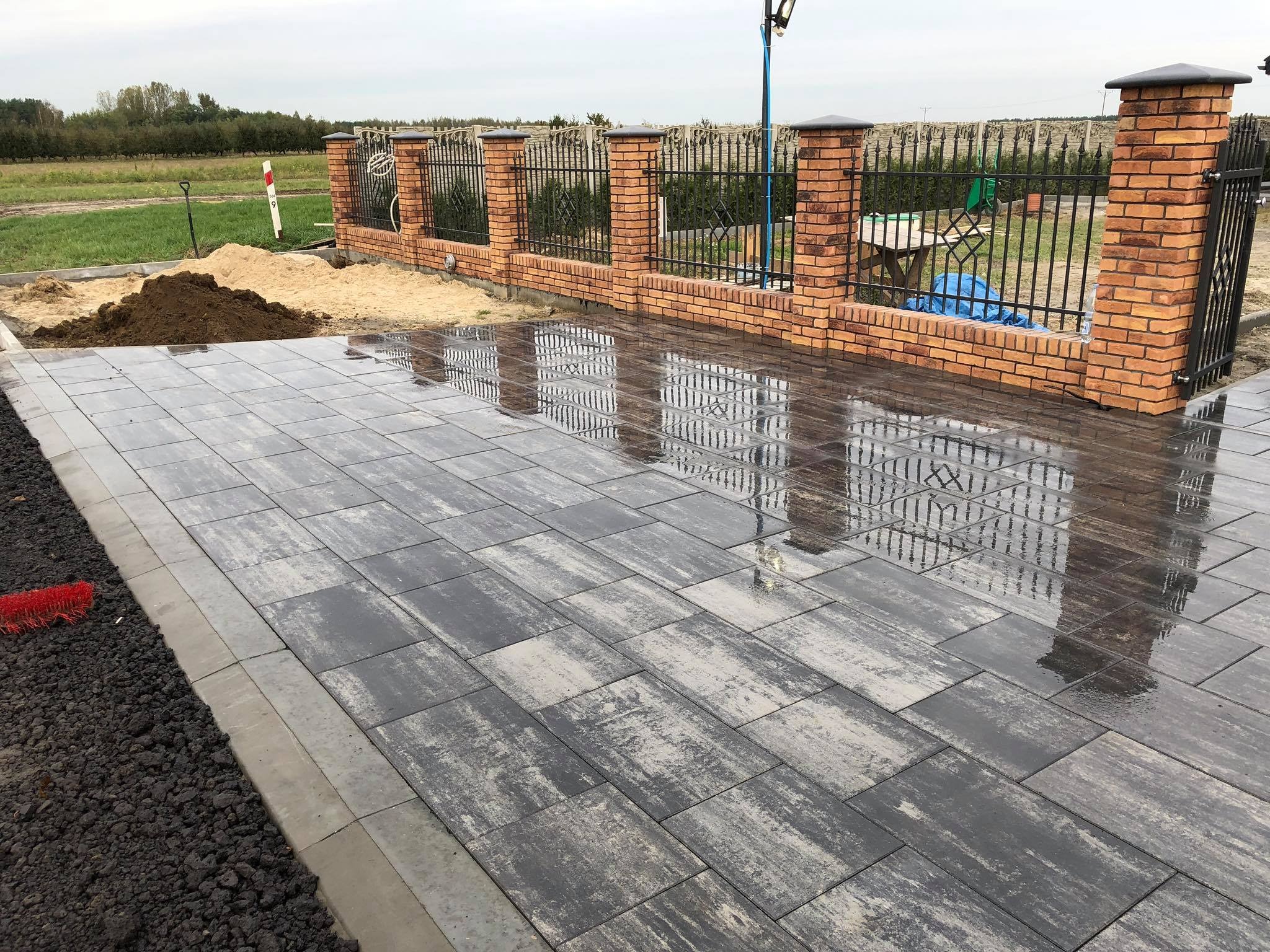 Świeżo ułożona, mokra nawierzchnia z prostokątnej kostki w odcieniach szarości, odbijająca w swojej powierzchni metalowe ogrodzenie z ceglanymi słupkami; w tle widoczna pryzma piasku i ziemi.