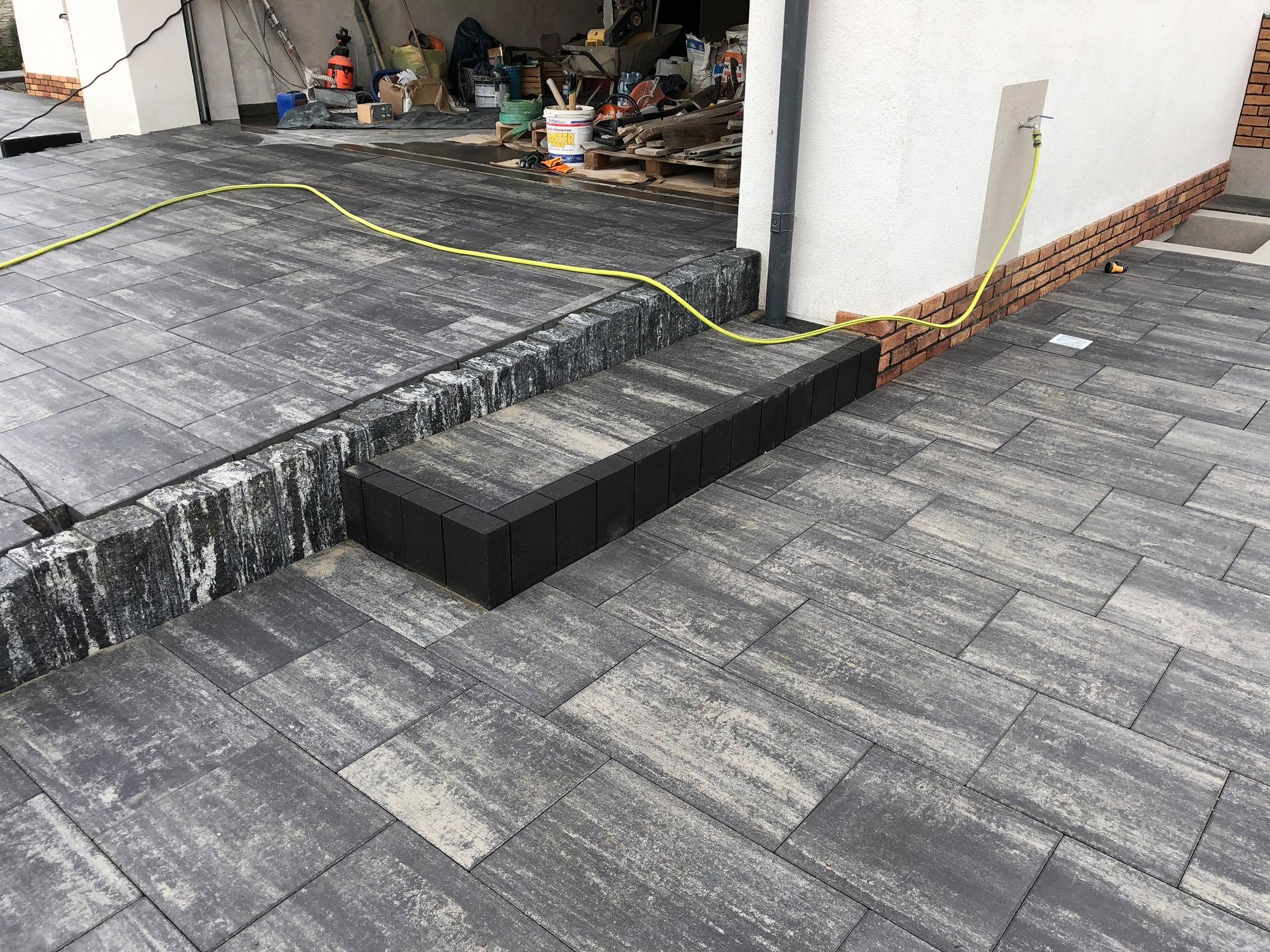 Ułożona nawierzchnia z kostki brukowej w odcieniach szarości, z widocznym fragmentem schodów wykończonych czarną kostką, żółty wąż ogrodowy leżący na powierzchni, w tle widoczne materiały budowlane.