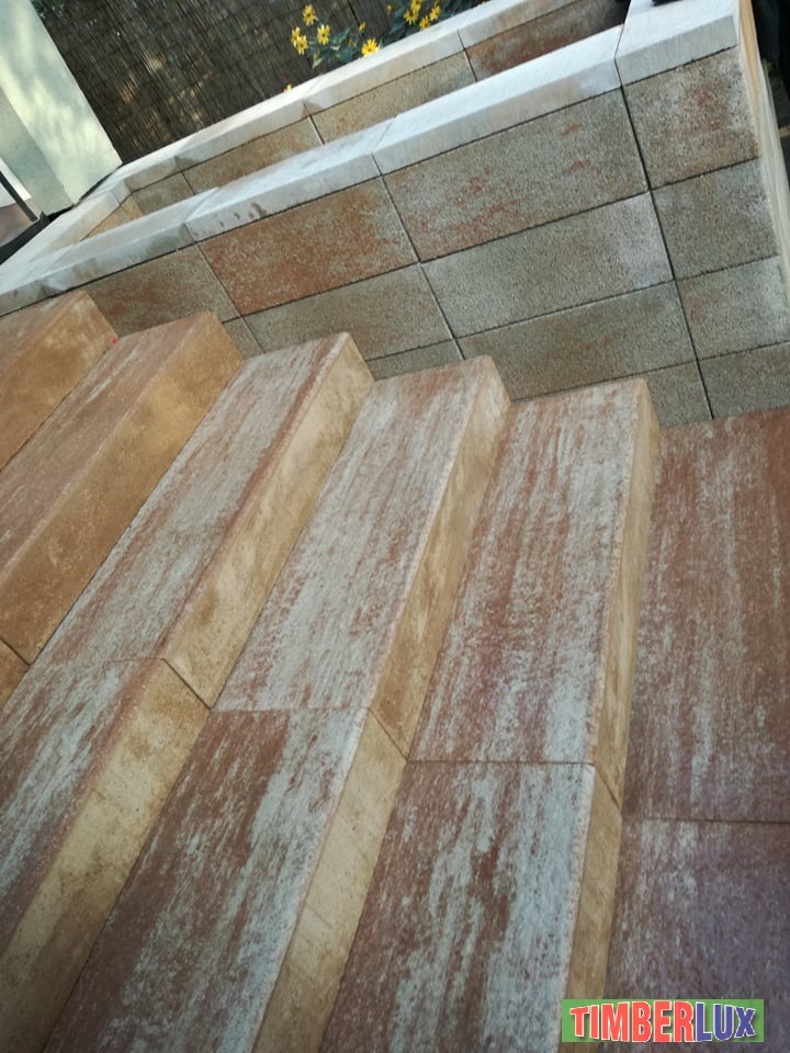 Schody zewnętrzne wyłożone brązowo-beżową kostką z efektem przetarć, prowadzące do podwyższonego klombu z jasnoszarą kostką, w tle bambusowa mata i żółte kwiaty.