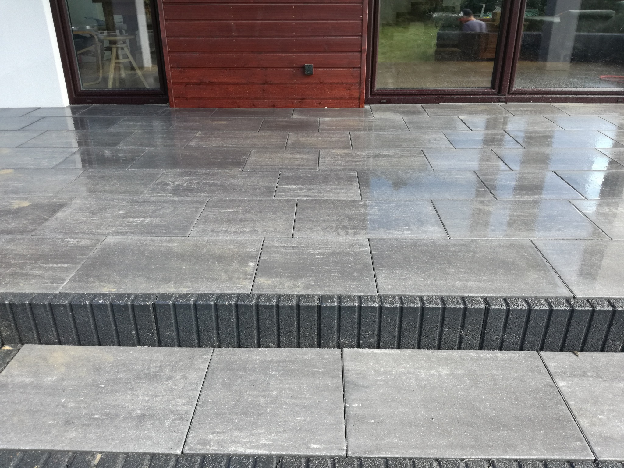 Świeżo ułożony chodnik z szarej kostki brukowej przed domem, z widocznymi stopniami wykończonymi czarnym obrzeżem. Powierzchnia kostki jest mokra, odbijająca światło.