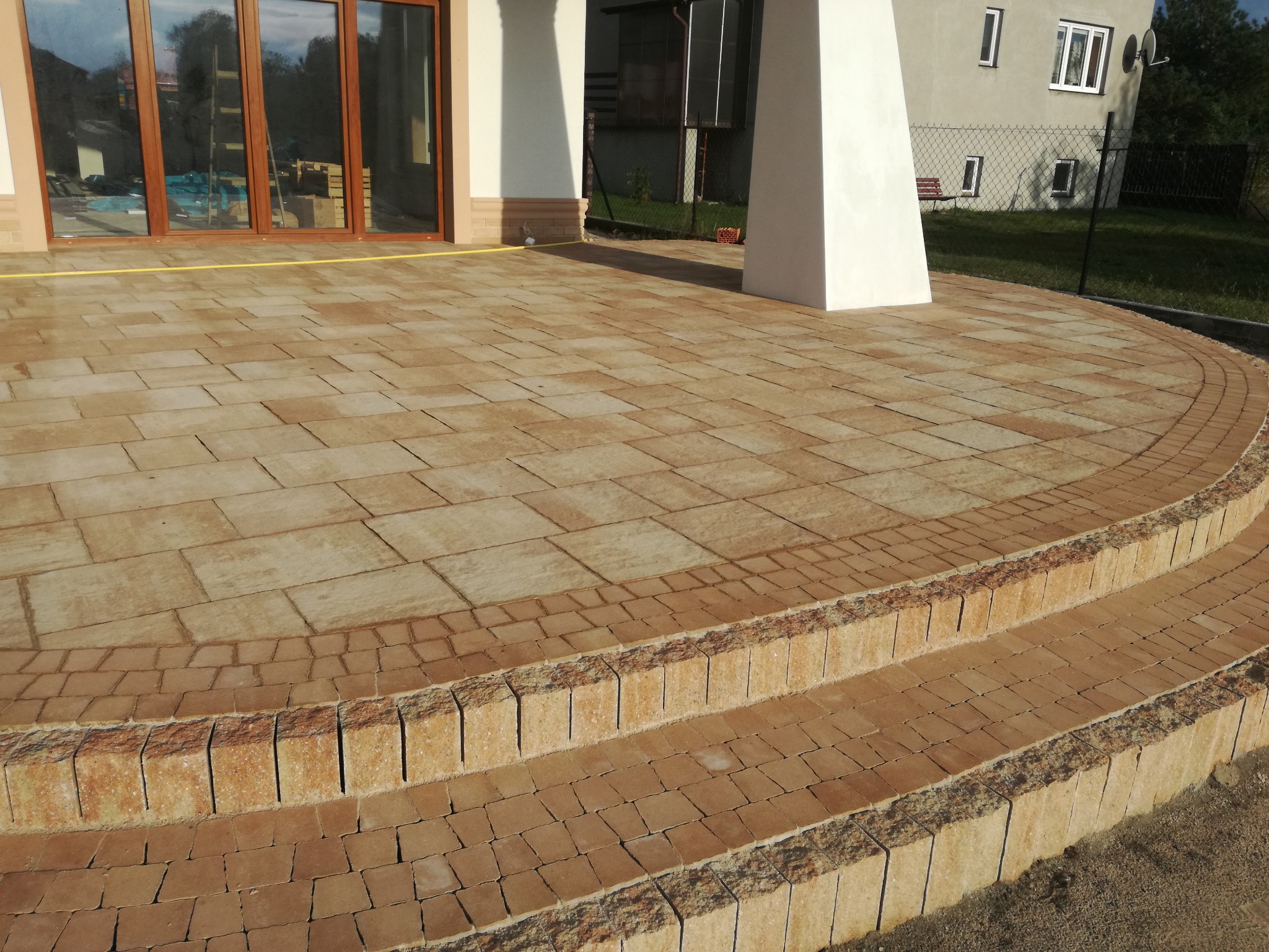 Taras wyłożony beżowo-brązową kostką brukową z dwoma stopniami, widok na budynek z dużymi oknami tarasowymi i białą kolumną.