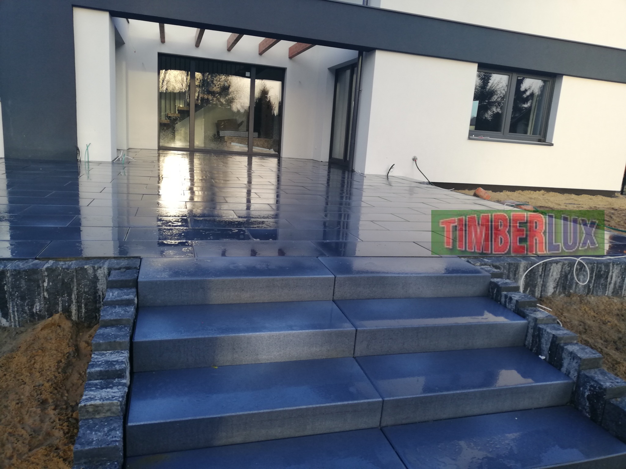 Nowoczesny taras z ciemnej kostki brukowej po deszczu, schody wykończone obrzeżami z kamienia łupanego, widok na dom z dużymi oknami.