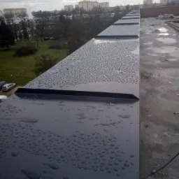 Metalowa obróbka blacharska na dachu pokryta kroplami deszczu, w tle budynki mieszkalne i pochmurne niebo.