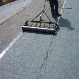 Prace dekarskie: aplikacja papy termozgrzewalnej na dachu płaskim za pomocą specjalistycznego wałka z palnikami, widoczne łączenia papy.