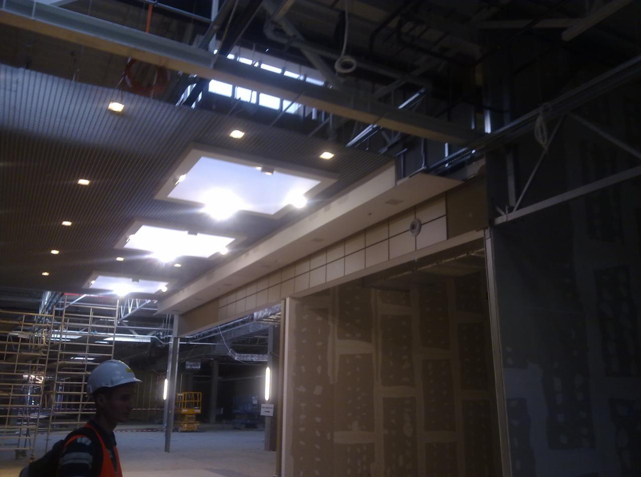 Montaż sufitu podwieszanego w CH FELICITY: widok na metalowy stelaż, oświetlenie w kasetonach i pracownika w kasku na tle rusztowań.