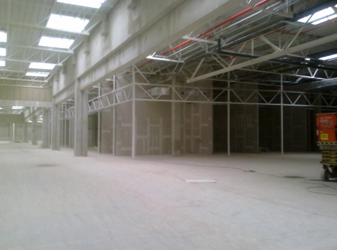 Wnętrze hali w trakcie budowy ścian działowych z widoczną konstrukcją szkieletową i częściowo zamontowanymi płytami. Widoczne stalowe słupy, elementy instalacji pod sufitem oraz podnośnik koszowy...