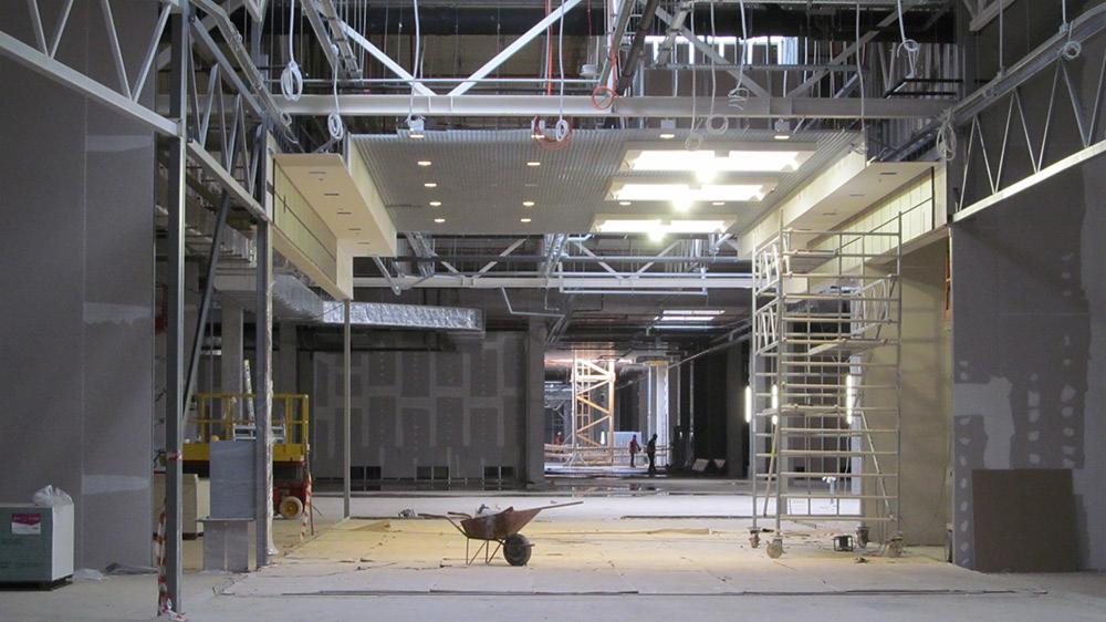 Wnętrze komercyjne w trakcie wykańczania: widoczne metalowe konstrukcje sufitowe, częściowo zamontowane panele oświetleniowe, ściany przygotowane do wykończenia, wózek budowlany i rusztowanie.