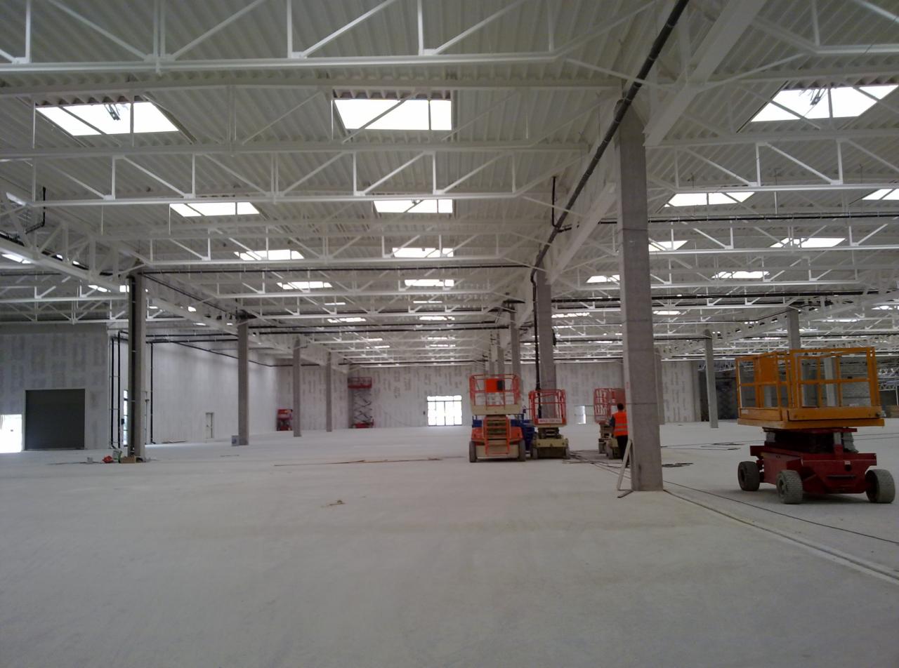 Wnętrze hali przemysłowej w trakcie prac wykończeniowych, widoczne surowe ściany, betonowa posadzka, stalowa konstrukcja dachu z oknami oraz podnośniki koszowe.
