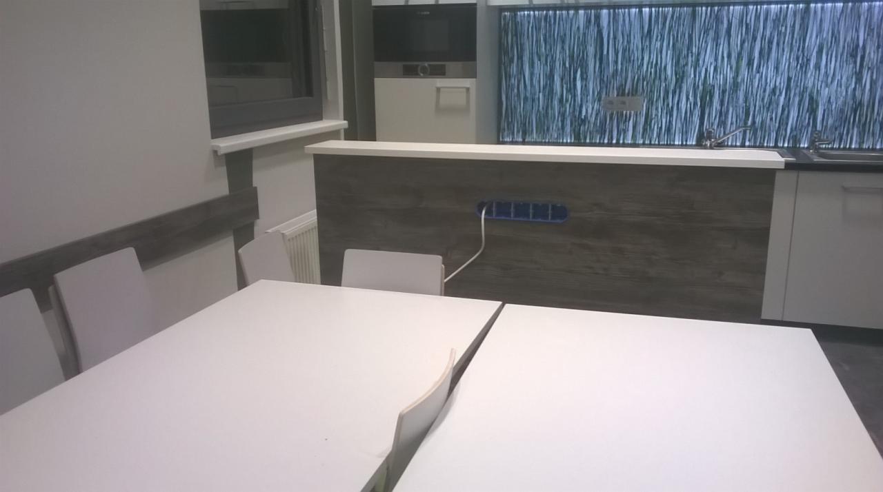 Nowoczesna aranżacja wnętrza z białym stołem, krzesłami i zabudową kuchenną z drewnopodobnym frontem oraz podświetlanym panelem dekoracyjnym imitującym trawę.