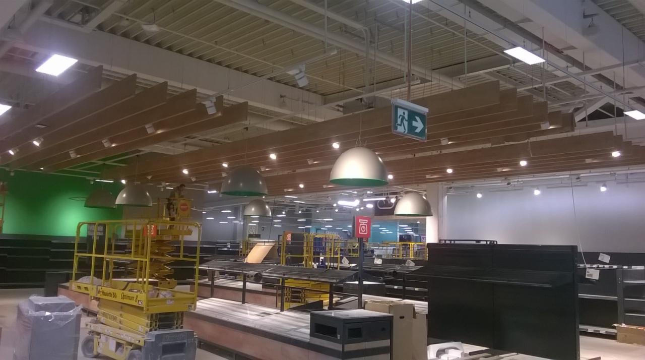 Montaż drewnianych elementów dekoracyjnych pod sufitem w supermarkecie Kaufland, widoczny podnośnik koszowy i oprawy oświetleniowe.