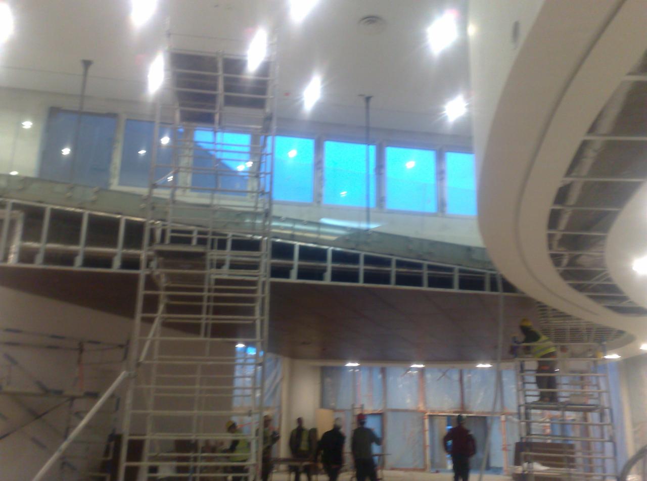 Wnętrze w trakcie wykończenia, widoczne rusztowania, pracownicy, okna z niebieską folią, oraz konstrukcja sufitu podwieszanego w kształcie łuku.