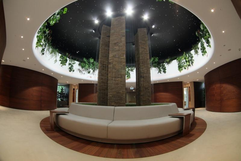 Okrągłe wnętrze hotelu Narvil z centralną instalacją: trzy kamienne kolumny, otoczone zielenią i okrągłą sofą. Sufit podwieszany z motywem gwiazd i roślinnością.