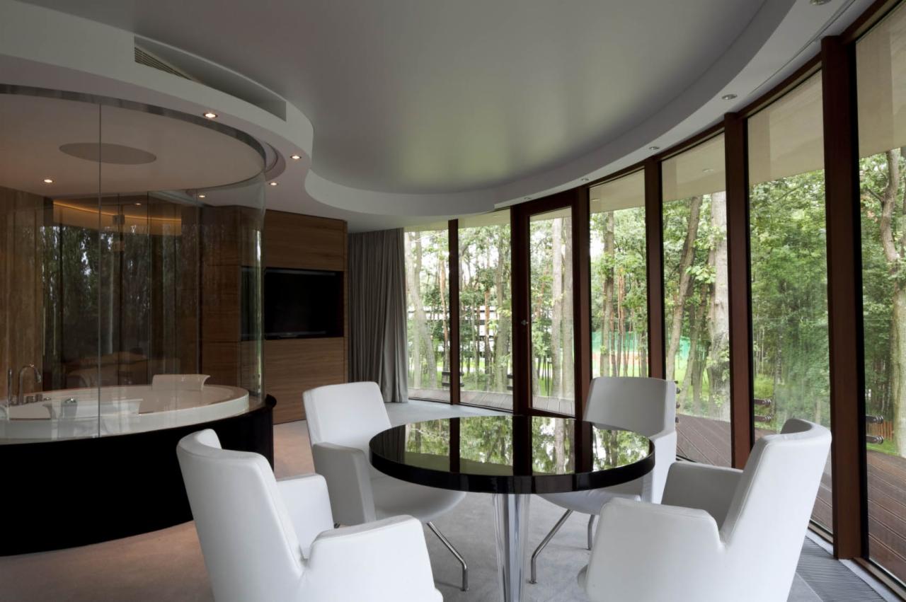 Wnętrze hotelu Narvil z okrągłym stołem z czarnym blatem i białymi krzesłami, widok na las przez panoramiczne okna, obok przeszklona łazienka z okrągłą wanną.