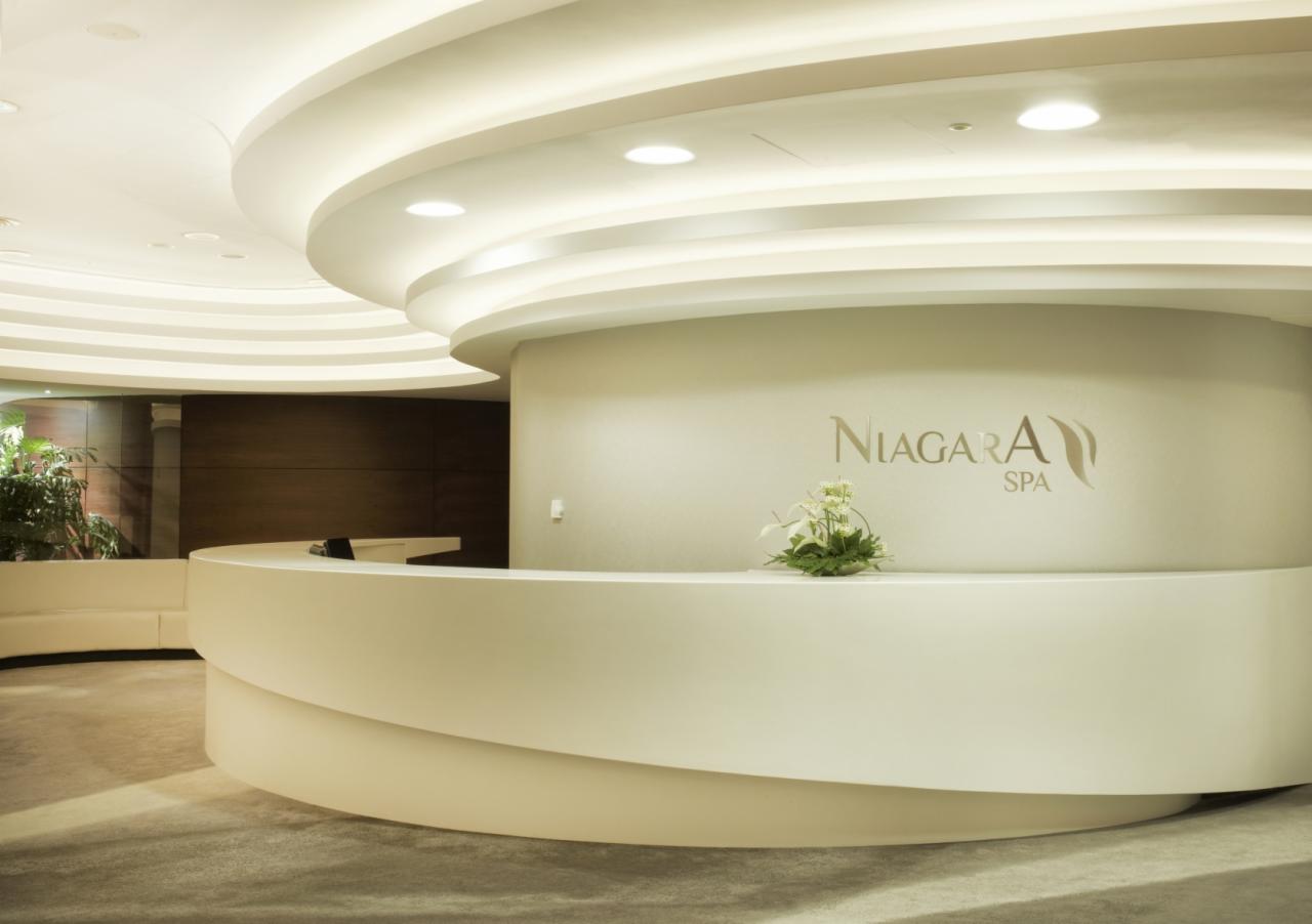 Nowoczesna recepcja hotelowa z zaokrąglonym blatem w kolorze złamanej bieli, logo 'Niagara SPA' na ścianie i dekoracją kwiatową. Widoczny fragment sufitu z podwieszanymi, okrągłymi elementami...
