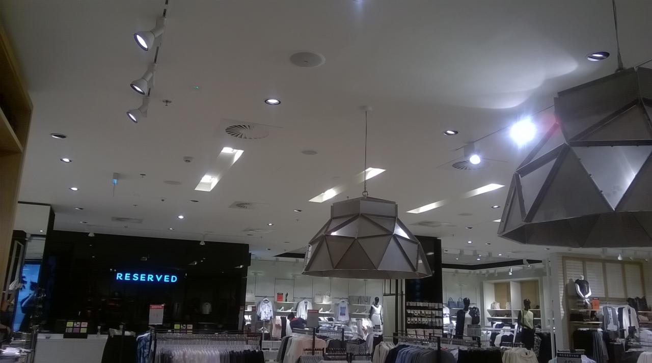 Wnętrze sklepu Reserved w CH Mokotów z widocznym oświetleniem sufitowym, w tym lampami wiszącymi o geometrycznym kształcie, reflektorami szynowymi i kratkami wentylacyjnymi.