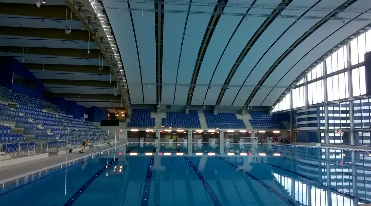 Kryty basen olimpijski w Lublinie z niebieskimi trybunami, sufitem z paneli i dużymi oknami odbijającymi się w wodzie, widok z poziomu wody.