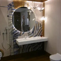 Nowoczesna łazienka z okrągłym lustrem z podświetleniem LED, białą umywalką nablatową, baterią i prysznicem ze szklaną kabiną, drewnianą podłogą i designerskimi lampami sufitowymi.