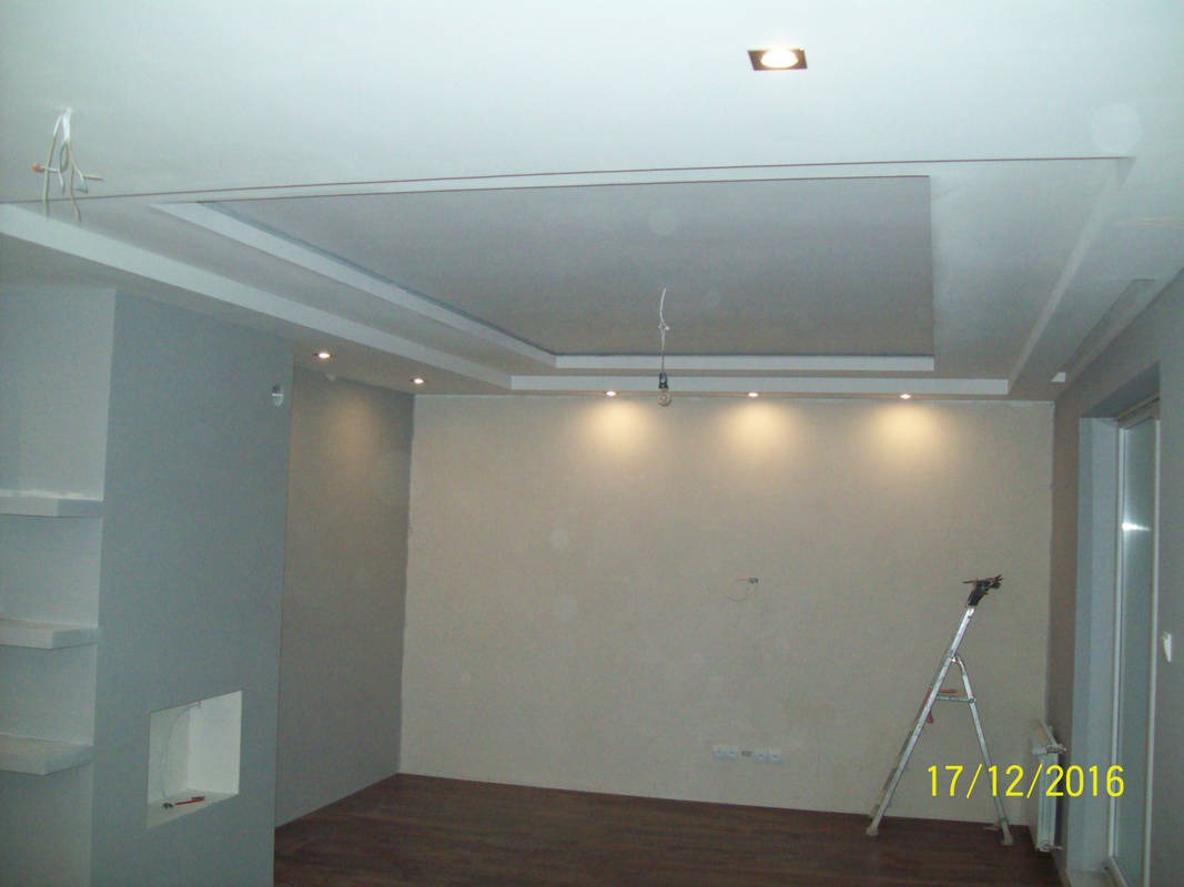 Wnętrze pokoju w trakcie prac wykończeniowych: widoczne gładzie na ścianach, podwieszany sufit z oświetleniem punktowym, drabina aluminiowa w rogu pomieszczenia, częściowo ułożona podłoga z paneli.